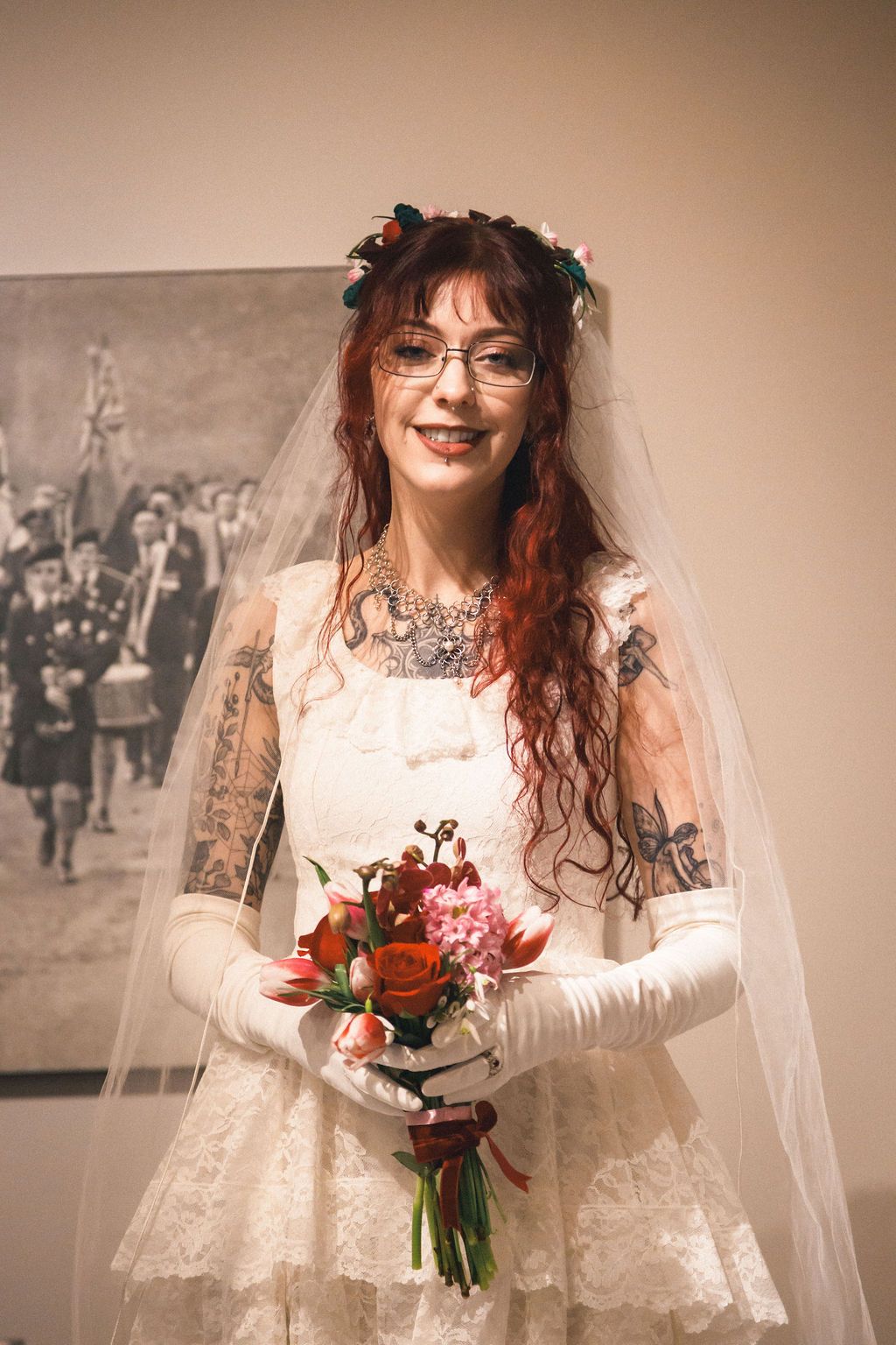 Tattooed bride with red hair, glasses, and veil smiling while holding colorful floral bouquet.