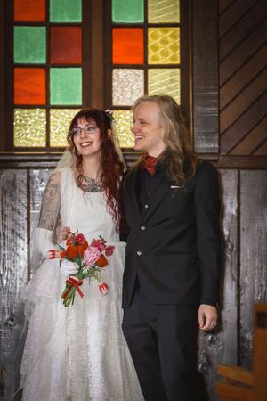 Joyful newlyweds smile together holding bridal bouquet before colorful stained glass chapel windows.