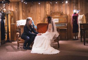 Bride in white lace gown sits with guest in rustic vintage-decorated wooden room.
