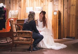 Tattooed bride in wedding gown laughs joyfully while chatting with guest in rustic interior.
