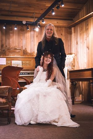 Bride in white lace gown sits with groom in black suit in a wood-paneled room with vintage organ.