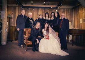 Wedding group portrait with bride in white gown and guests in formal attire in a rustic wood-paneled venue.