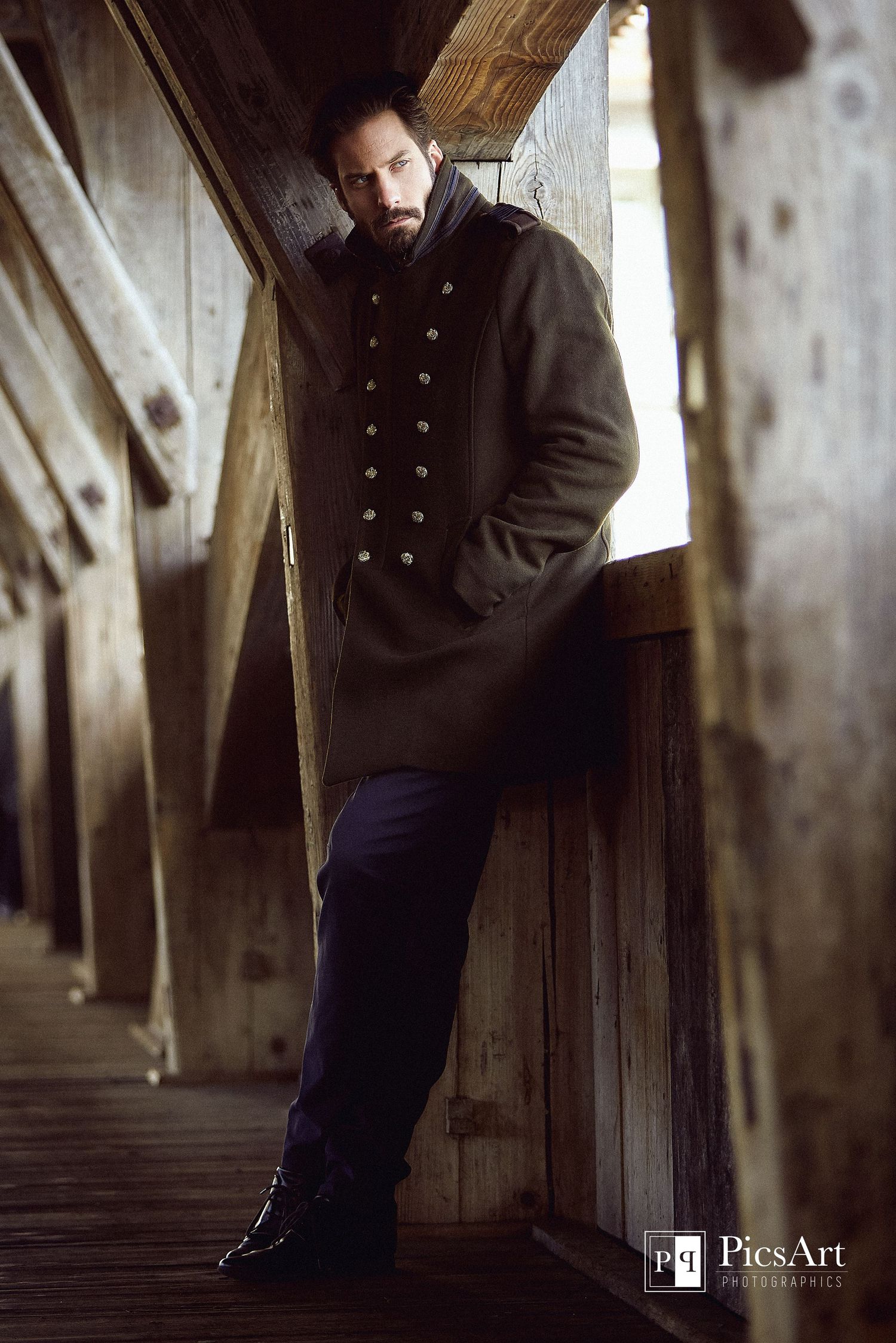 Male model in military-style coat leaning against wooden beams in moody portrait photography.