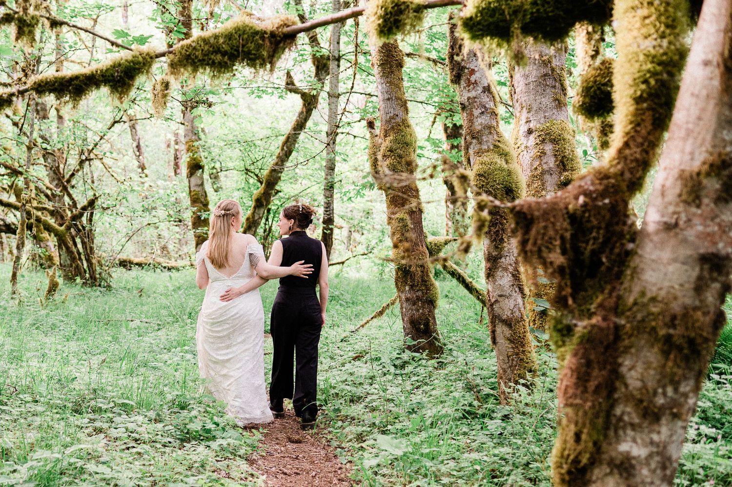 Two brides walk hand in hand through a lush forest setting surrounded by tall moss covered trees