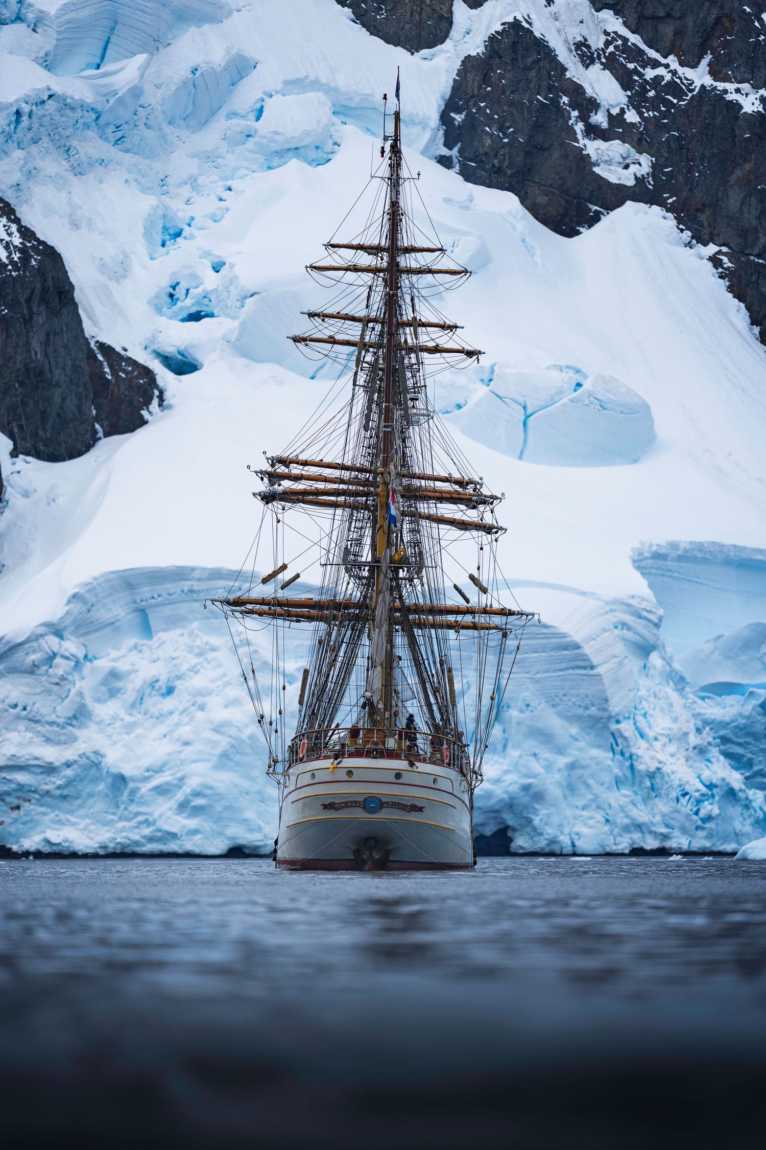 A tall sailing ship anchored in front of towering glaciers and icy cliffs in Antarctica, creating a dramatic polar seascape.