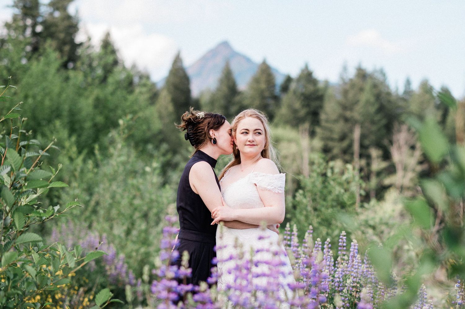 A couple embraces in a field of purple lupine wildflowers with evergreen trees and mountains in the background.