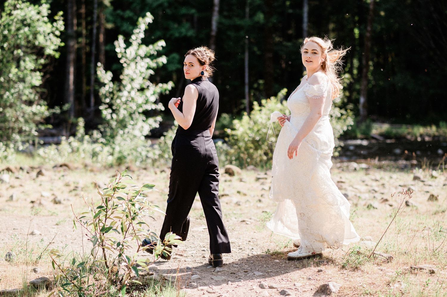 A couple in wedding attire walks together in a sunlit outdoor forest setting.
