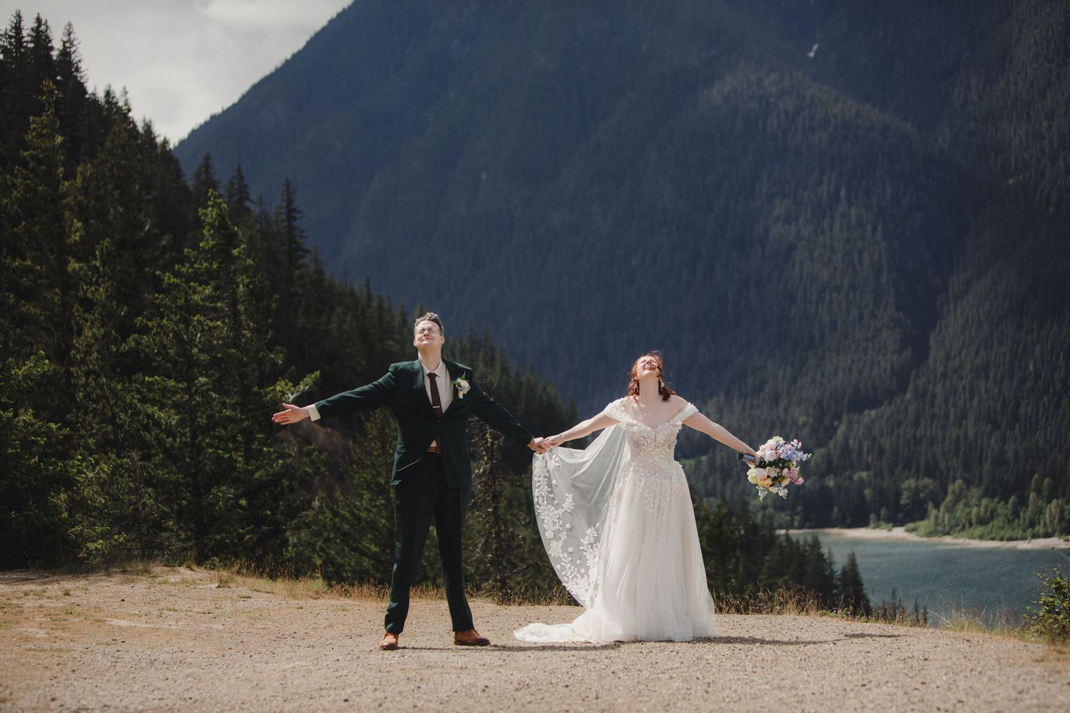 A couple poses for wedding photos against a dramatic mountain backdrop with evergreen trees and a scenic lake.