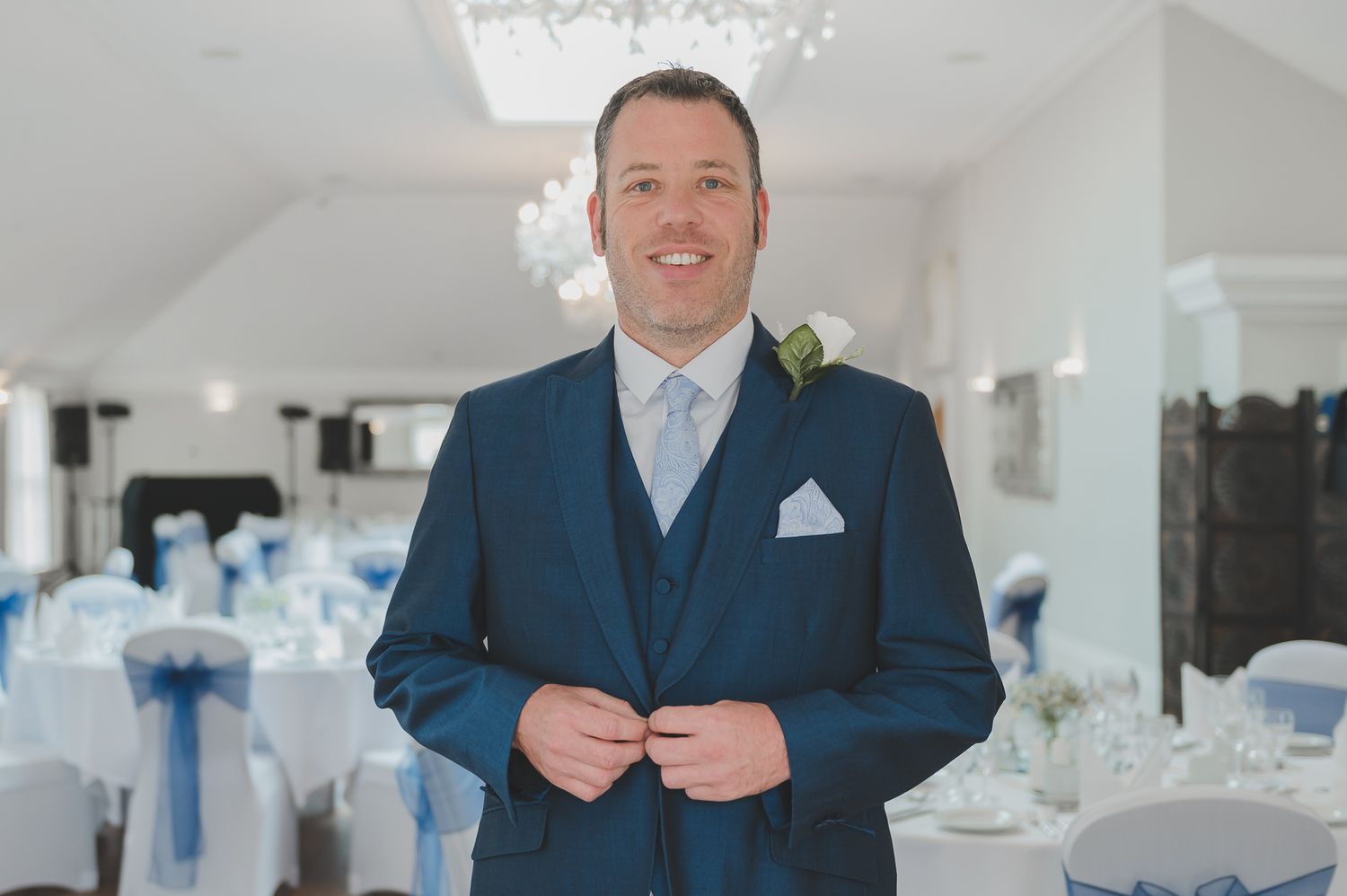 A groom in a navy blue suit and light blue tie stands smiling in an elegant wedding reception venue with blue decor.