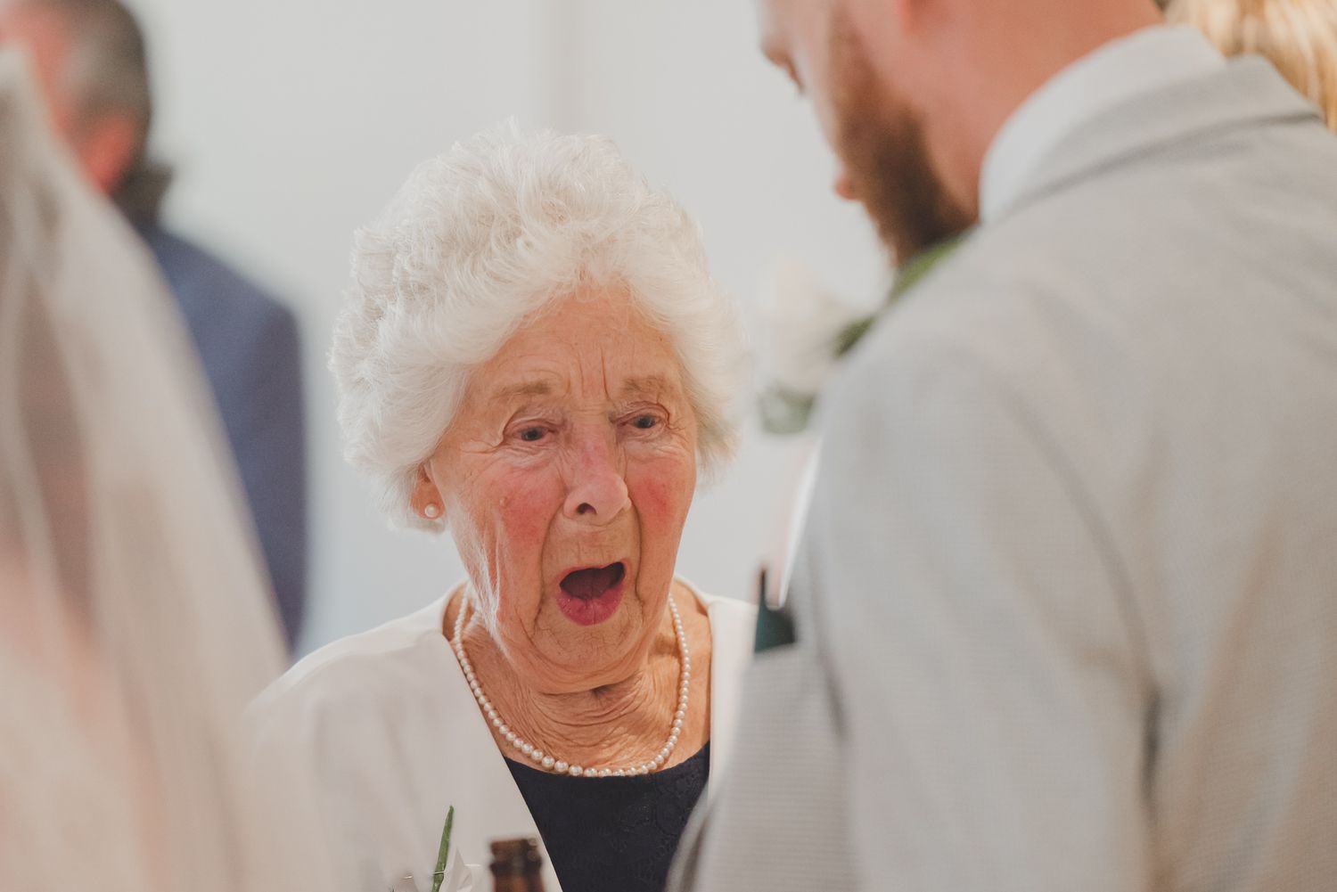 An elderly woman with white curly hair and pearl necklace reacts with surprise at a social gathering.