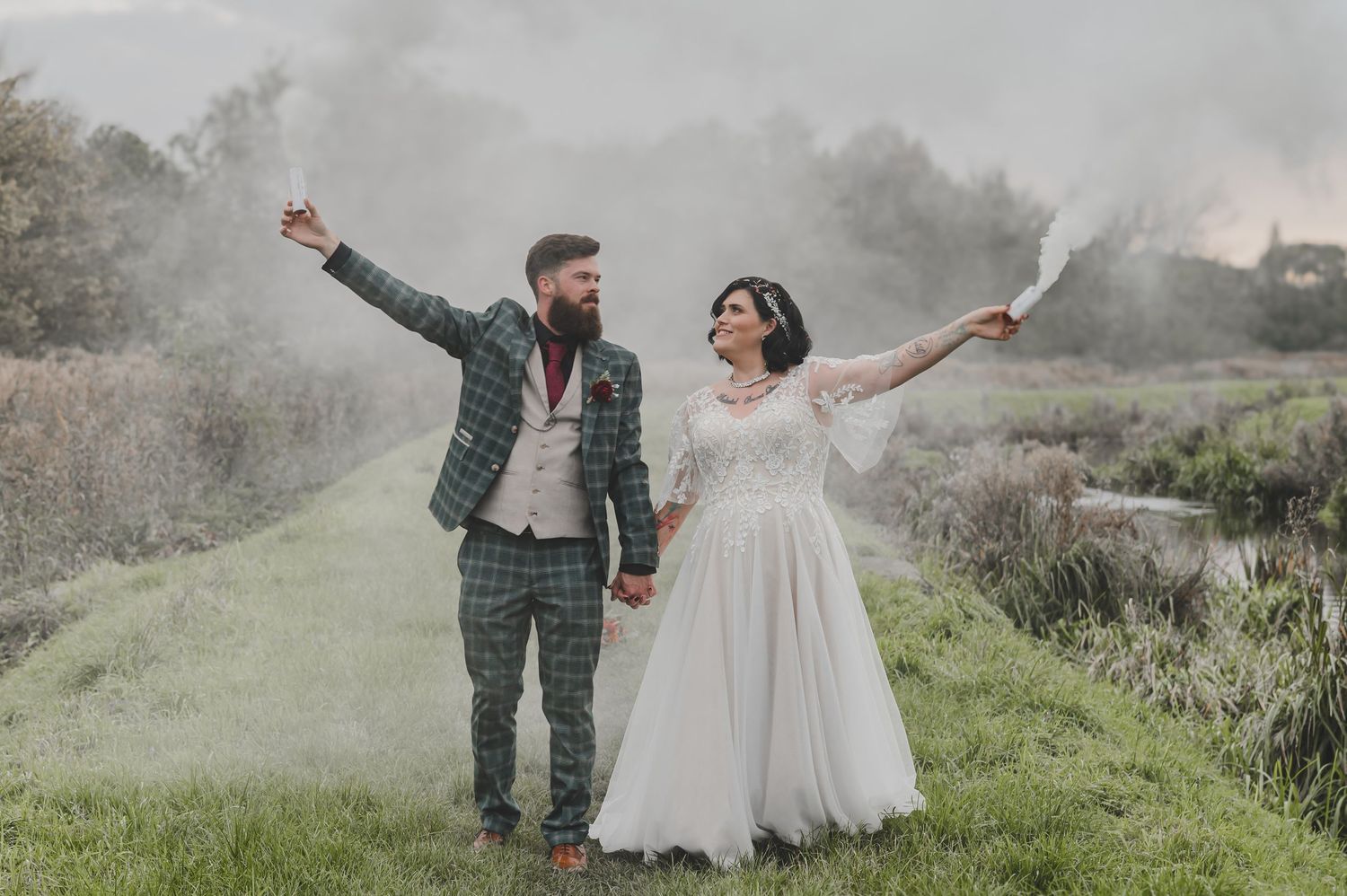 A couple in wedding attire holds hands while walking through a misty meadow at sunset.