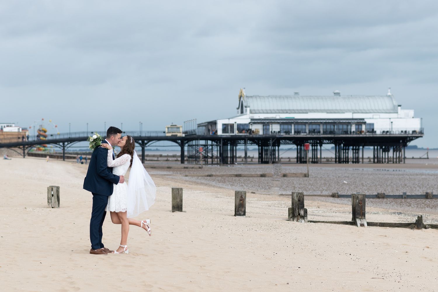 A romantic couple shares a kiss on a sandy beach with a wooden pier and cloudy sky in the background.