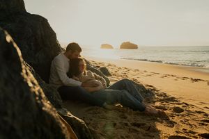 A couple sits together on a scenic beach at sunset with rocky cliffs in the background.