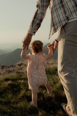 Adult and small child holding hands while walking on a grassy hillside at sunset.