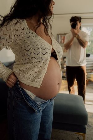 Person wearing white knit sweater and jeans showing pregnancy belly in home setting.