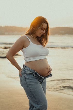 Person in white tank top and jeans showing pregnancy belly on beach at sunset.