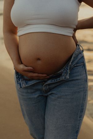 Person in white top and jeans showing pregnancy progress in beach setting at sunset.