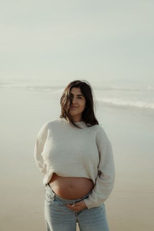 A person in a cream sweater poses on a beach during an overcast day.