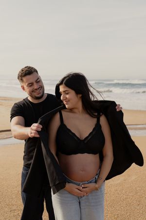 A couple shares playful moments on a sandy beach during a maternity photoshoot.