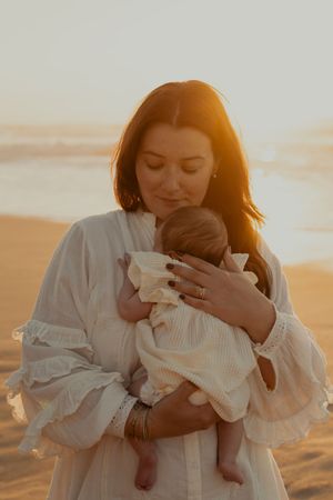 Mother in white dress holding baby during golden hour on beach.