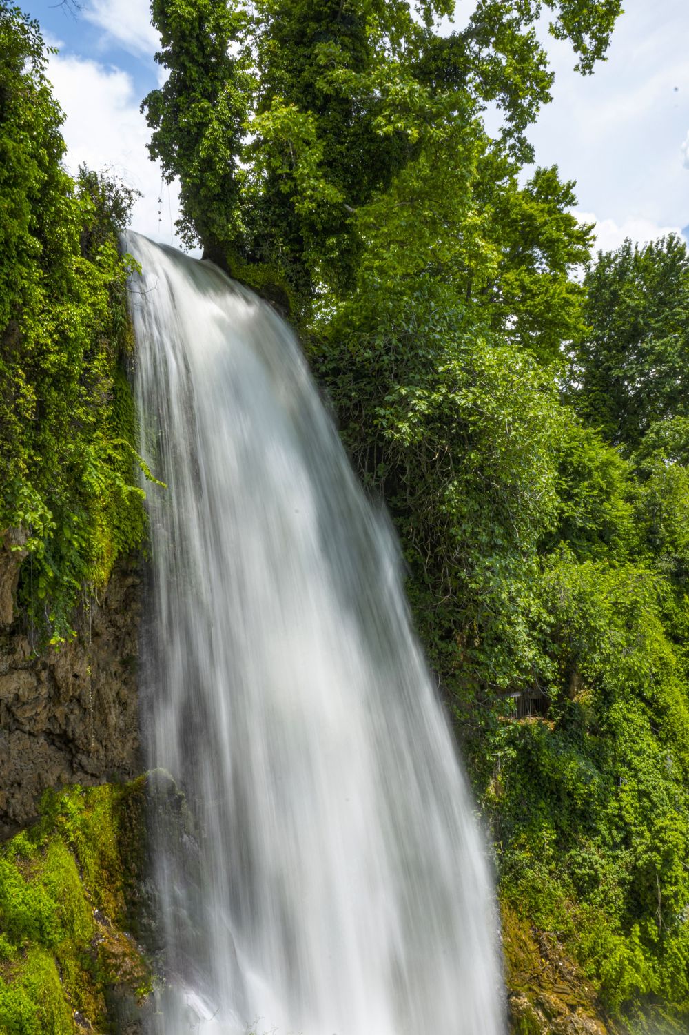 A tall waterfall cascades down a rocky cliff surrounded by lush green forest and trees on a sunny day.