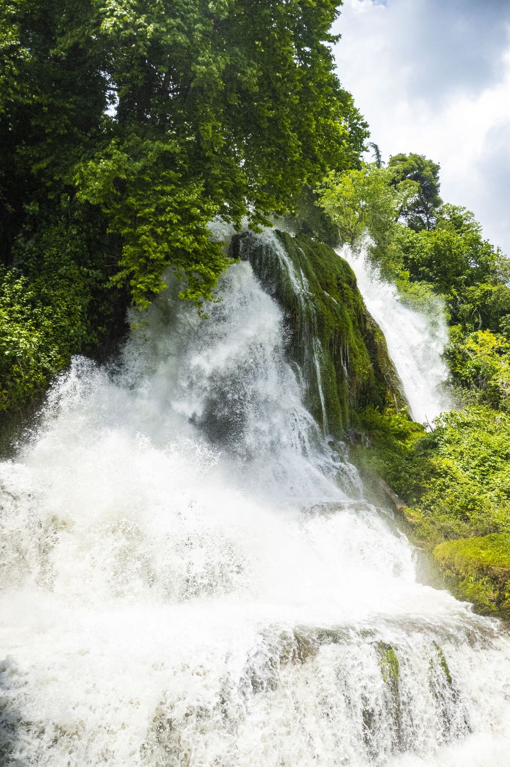 A powerful cascade of white water rushes down a rocky waterfall surrounded by lush green foliage on a cloudy day.