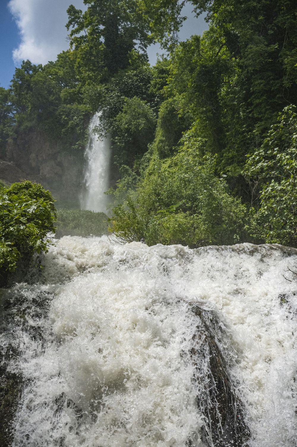 Powerful rushing waterfall cascades over rocks through lush green forest on a bright summer day.