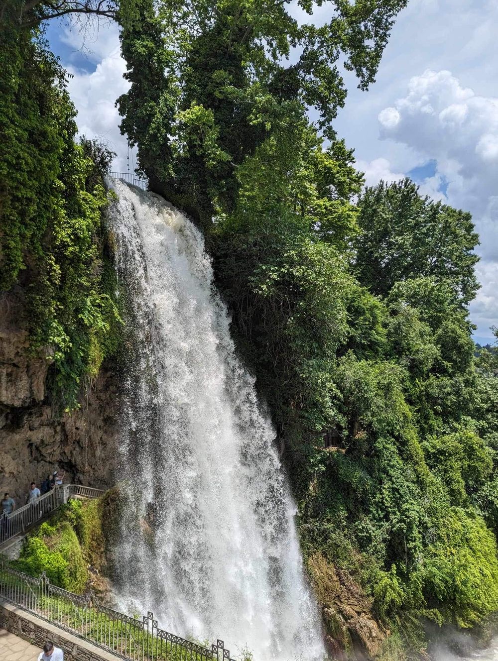 A majestic waterfall cascades down a rocky cliff surrounded by lush green foliage on a bright summer day.