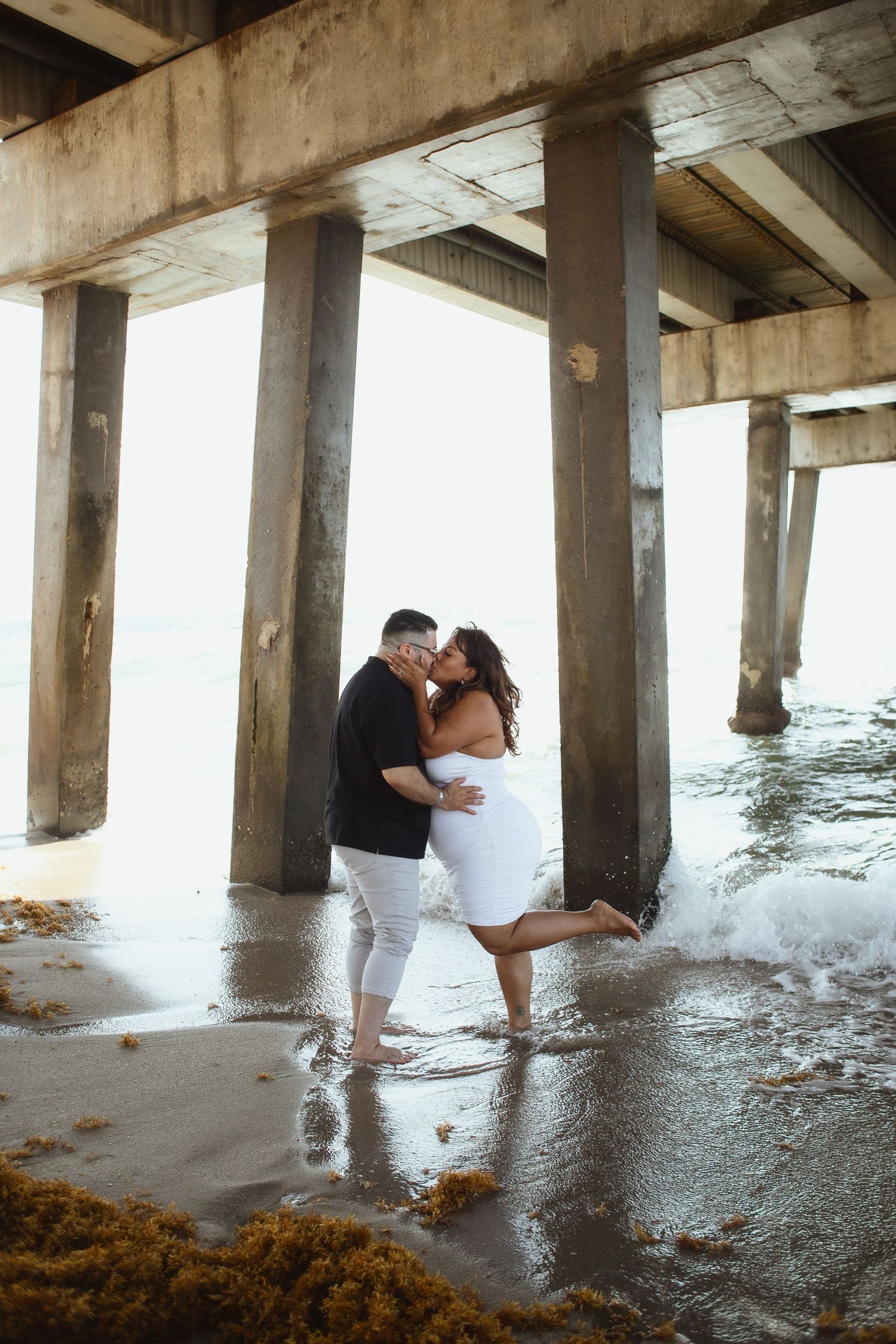 Couple kissing under Pompano Beach Pier during engagement couples session.