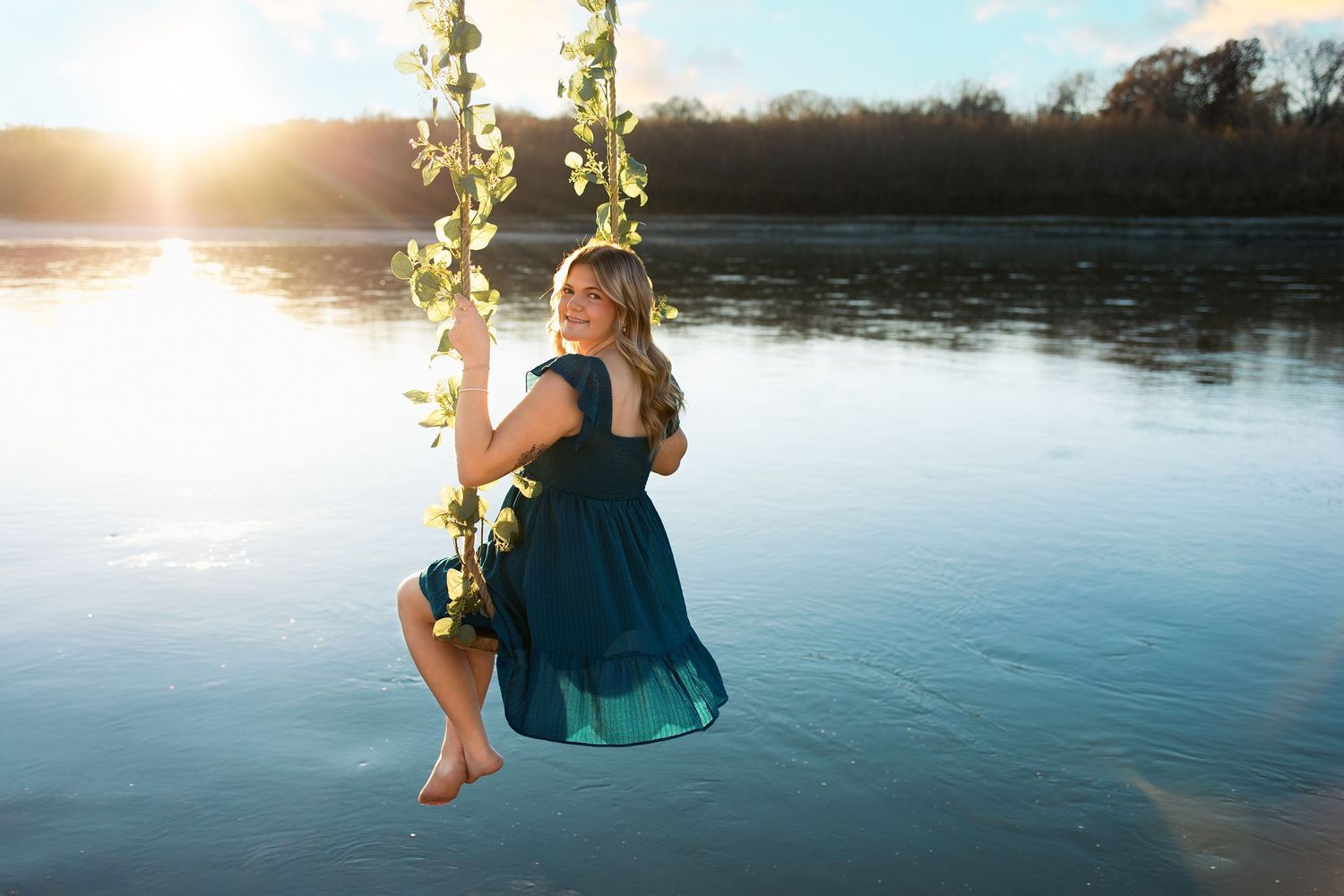 A senior girl swings on a rope swing over a tranquil lake at sunset, creating a peaceful summer scene.