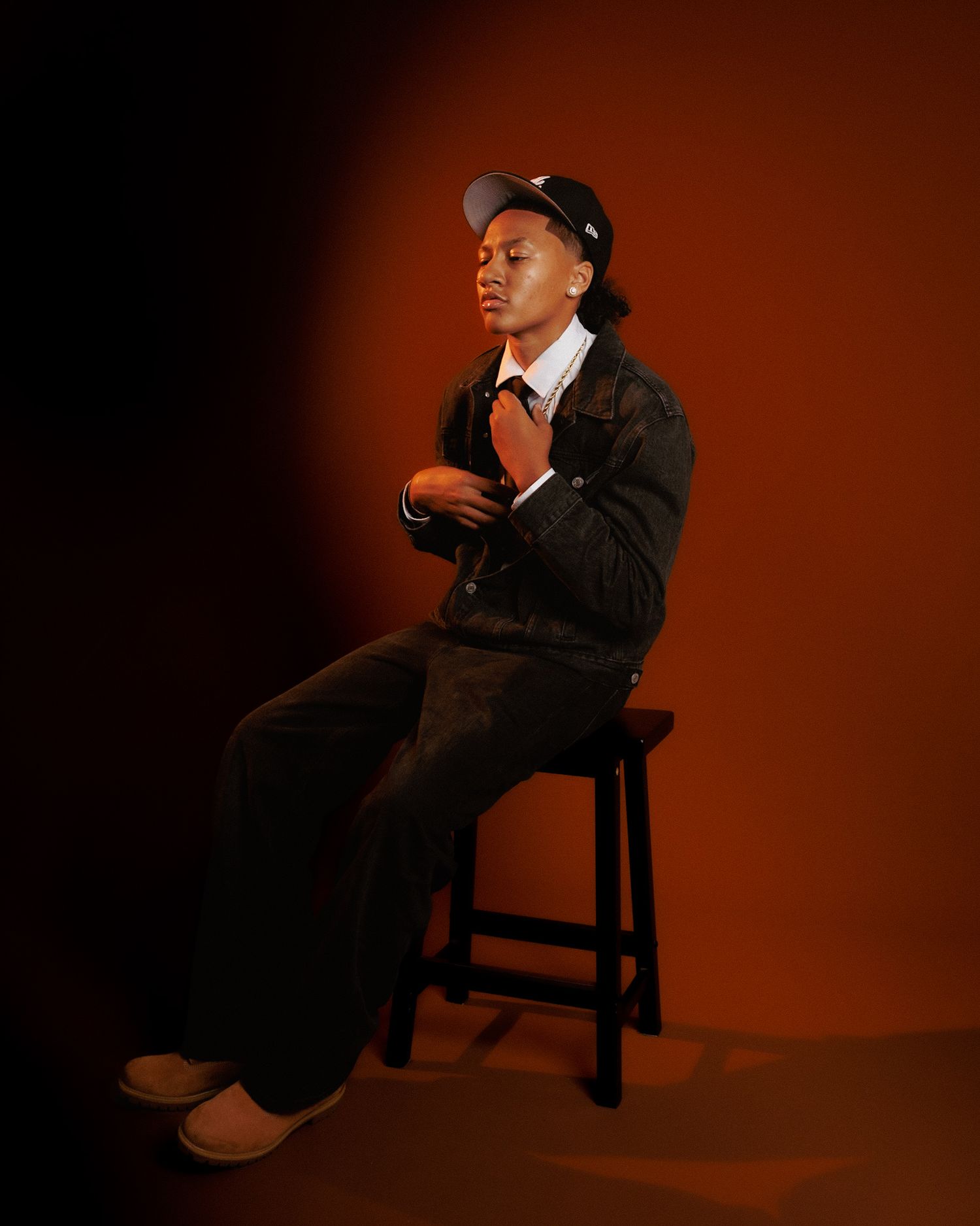 Figure in dark attire sits on wooden chair against reddish-brown backdrop in dramatic lighting.