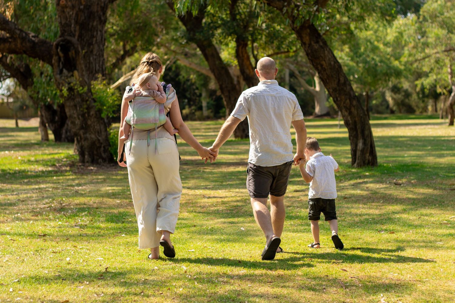Family walking hand-in-hand through a sunny green park, parents carrying and guiding young children among tall trees.