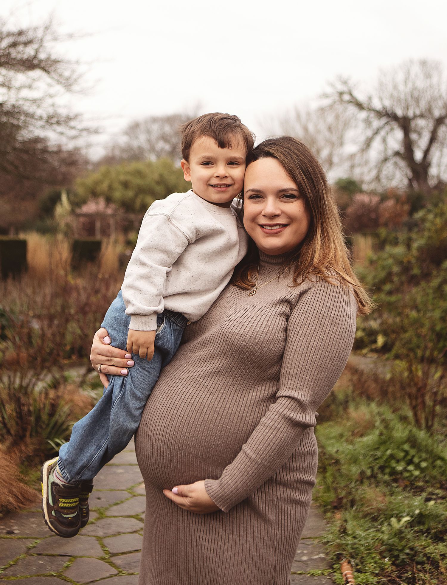 A person in a brown sweater playfully carries someone in their arms outdoors on a cloudy day. During a location maternity session in Bexley