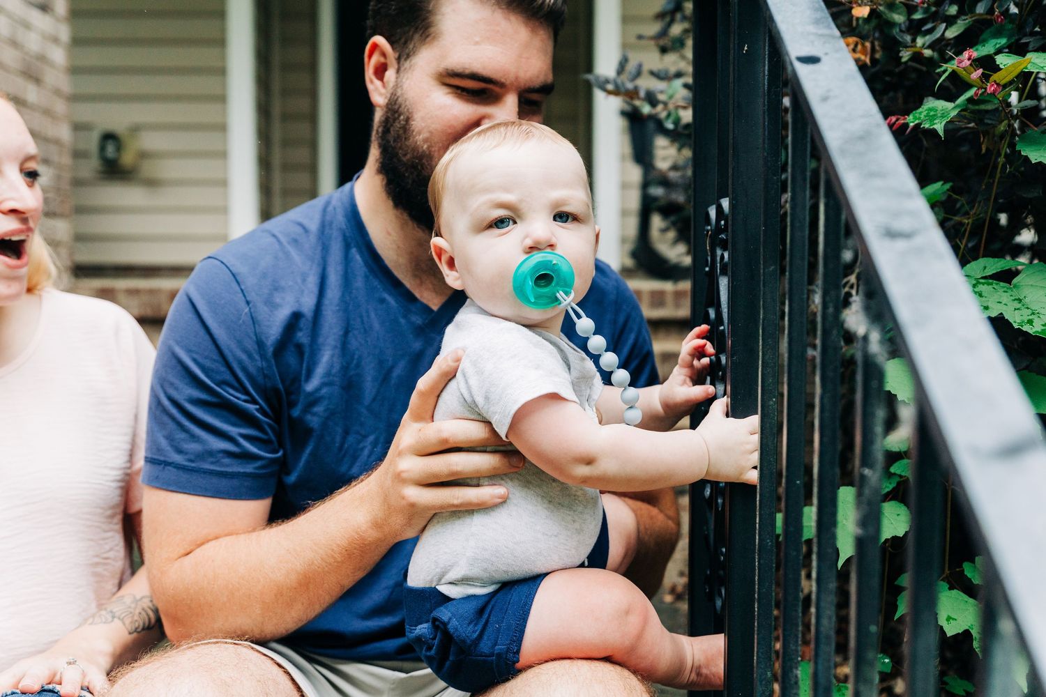 A family sits together on their front porch steps sharing a tender moment with their baby wearing a teal pacifier.