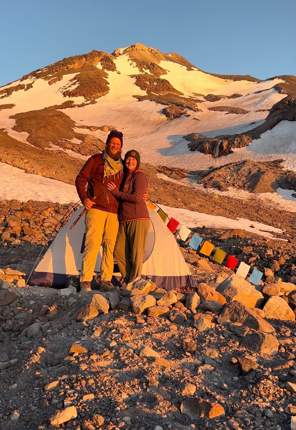Adventurous campers celebrate at sunset next to their tent on Mount Baker with snow-capped peaks in the background.