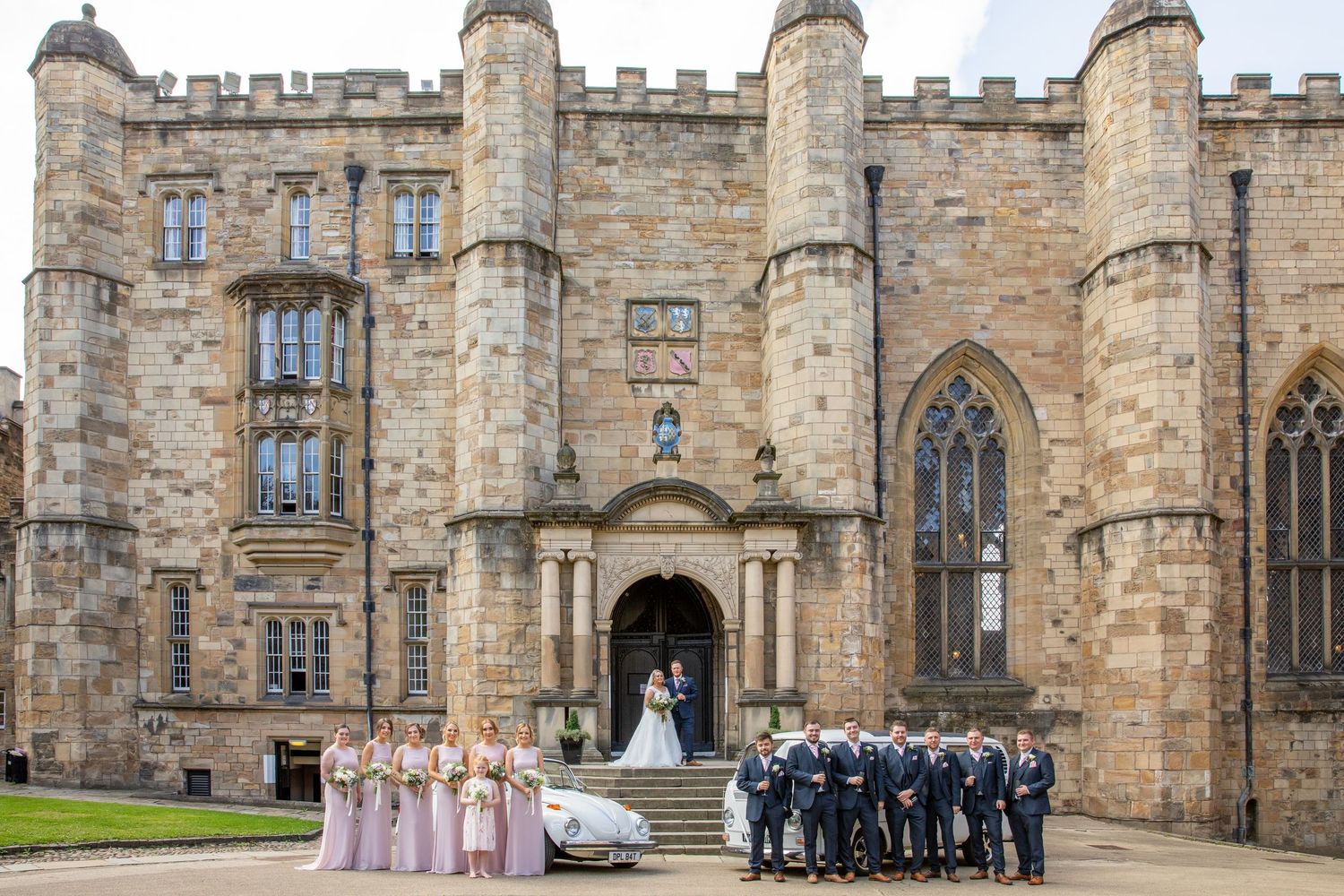 Wedding party gathers on the steps of Durham Castle's historic stone entrance with bridesmaids in pink and groomsmen in navy.