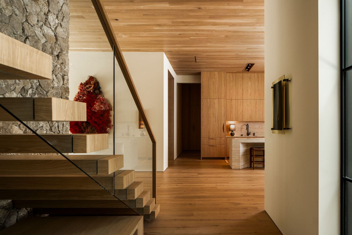 Modern interior hallway with wooden flooring, stone wall, floating staircase, and glass railing in warm lighting.