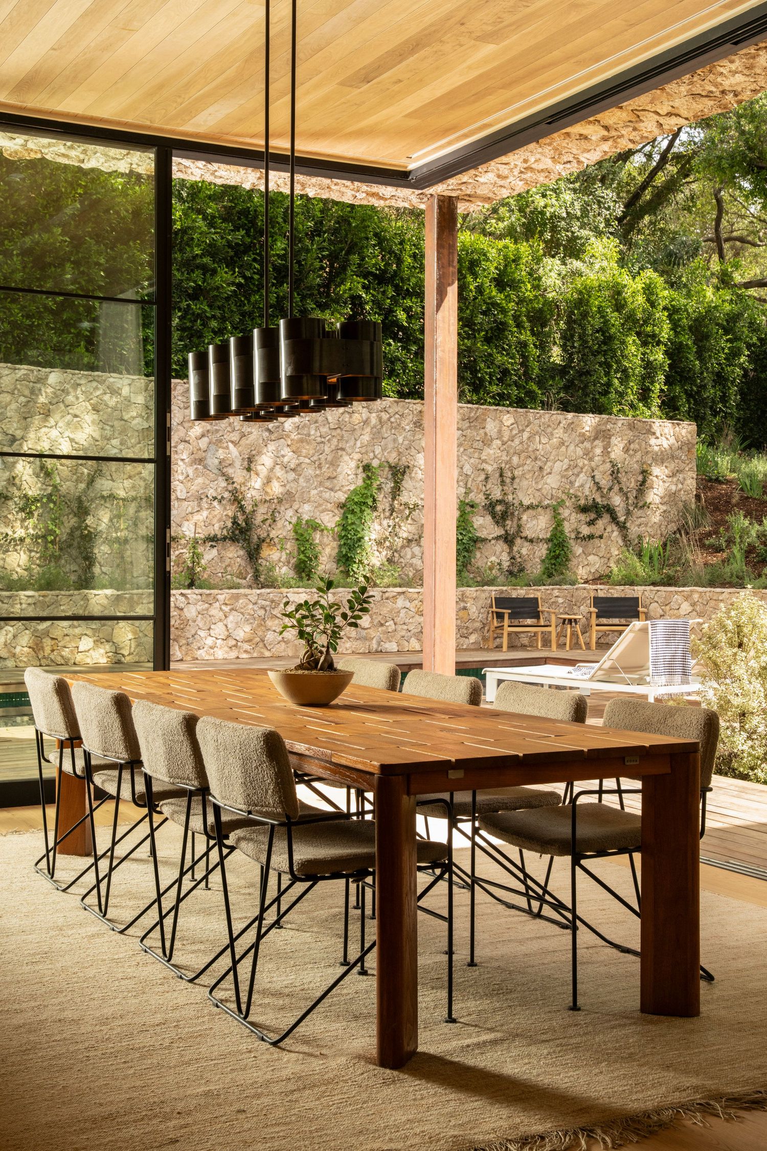 Modern outdoor dining area with wooden table and black chairs under a covered patio with stone wall backdrop.