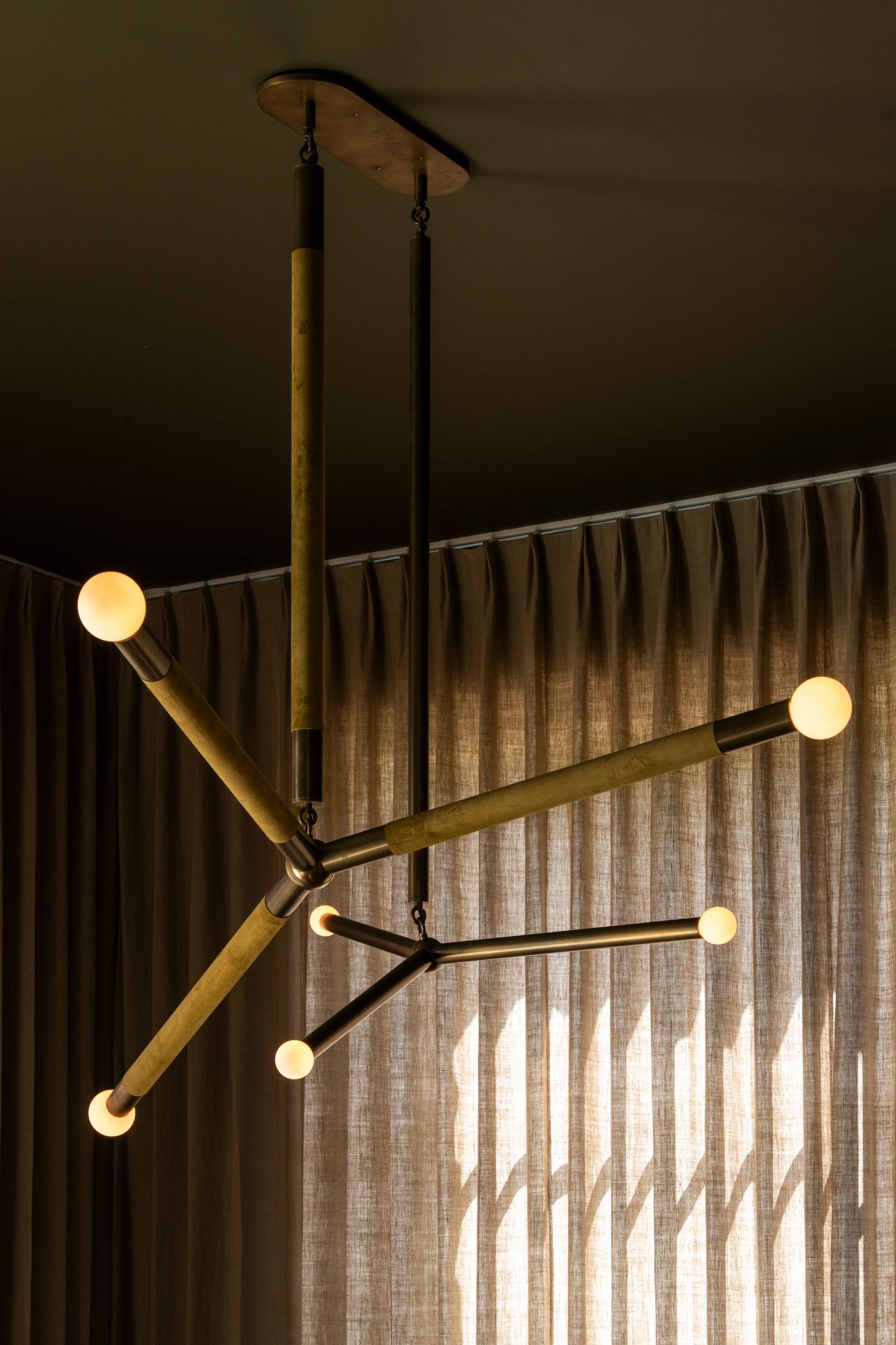 Modern geometric pendant light fixture with brass-toned arms and glowing spherical bulbs hangs against textured curtains.