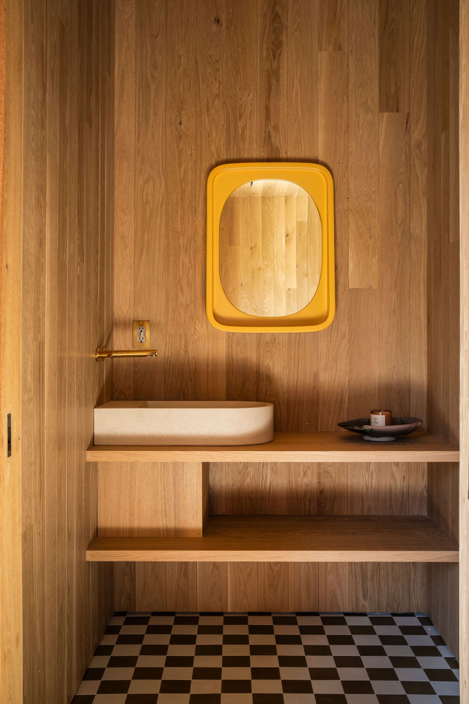 Modern bathroom with wood-paneled walls, yellow framed mirror, vessel sink, and checkered floor tiles.