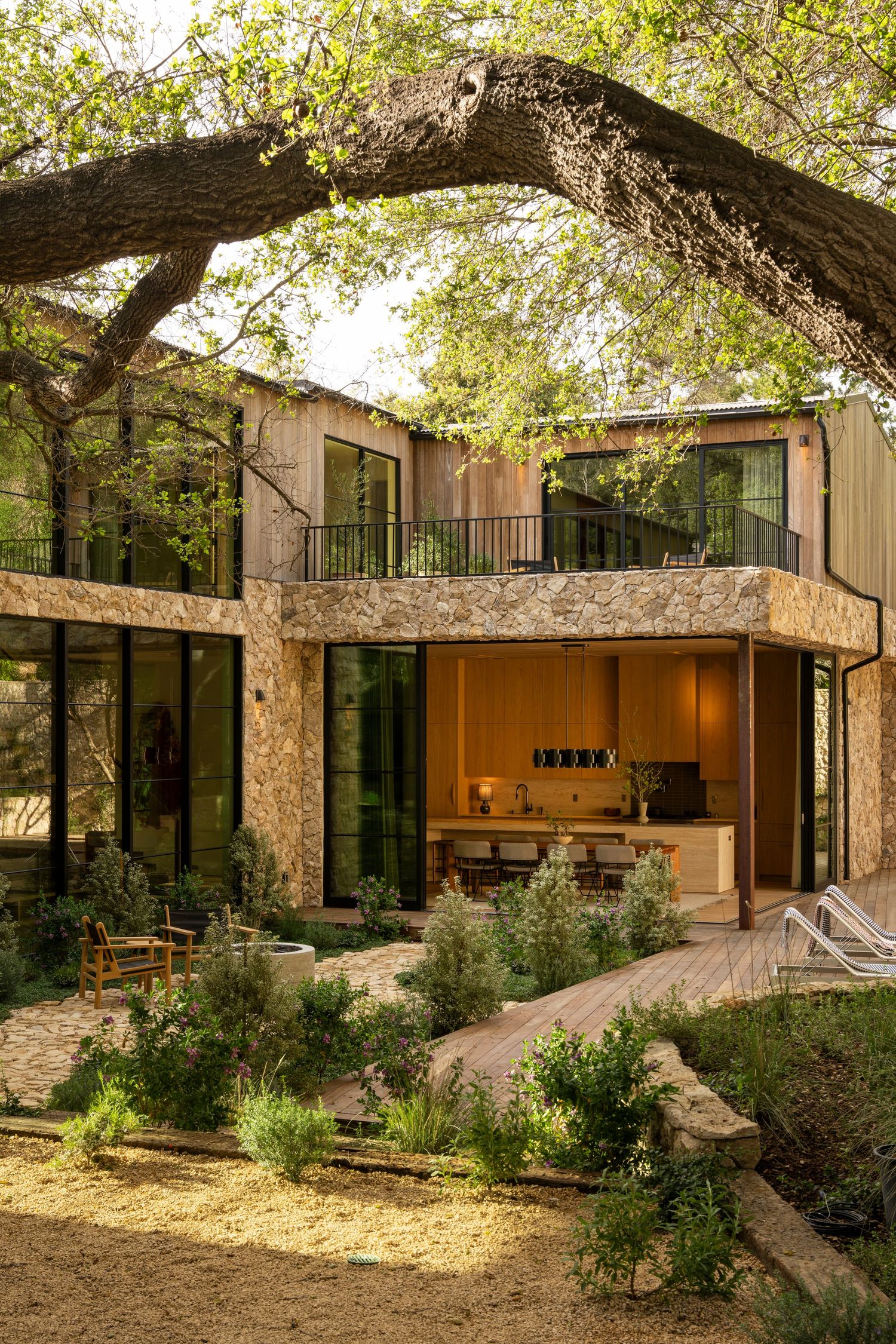 Modern stone and glass house with large windows and outdoor living space framed by overhanging tree branches in natural setting.