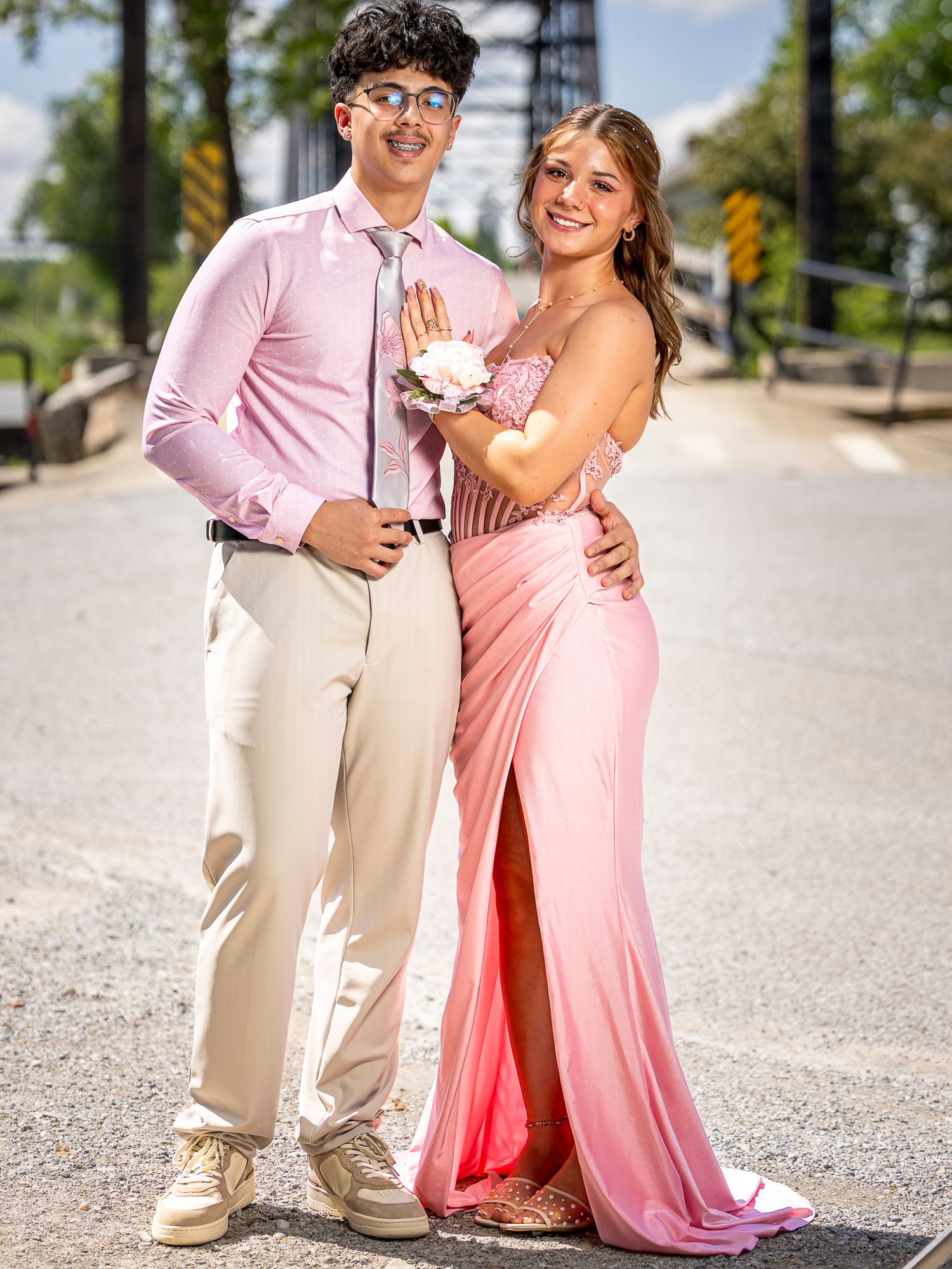 A couple dressed in coordinating pink prom attire poses together on a bridge, smiling for a formal photo.