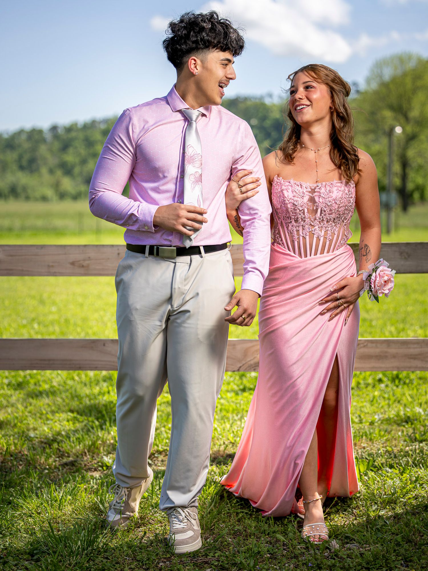 A young couple dressed in pink prom attire poses outdoors by a wooden fence on a sunny day.