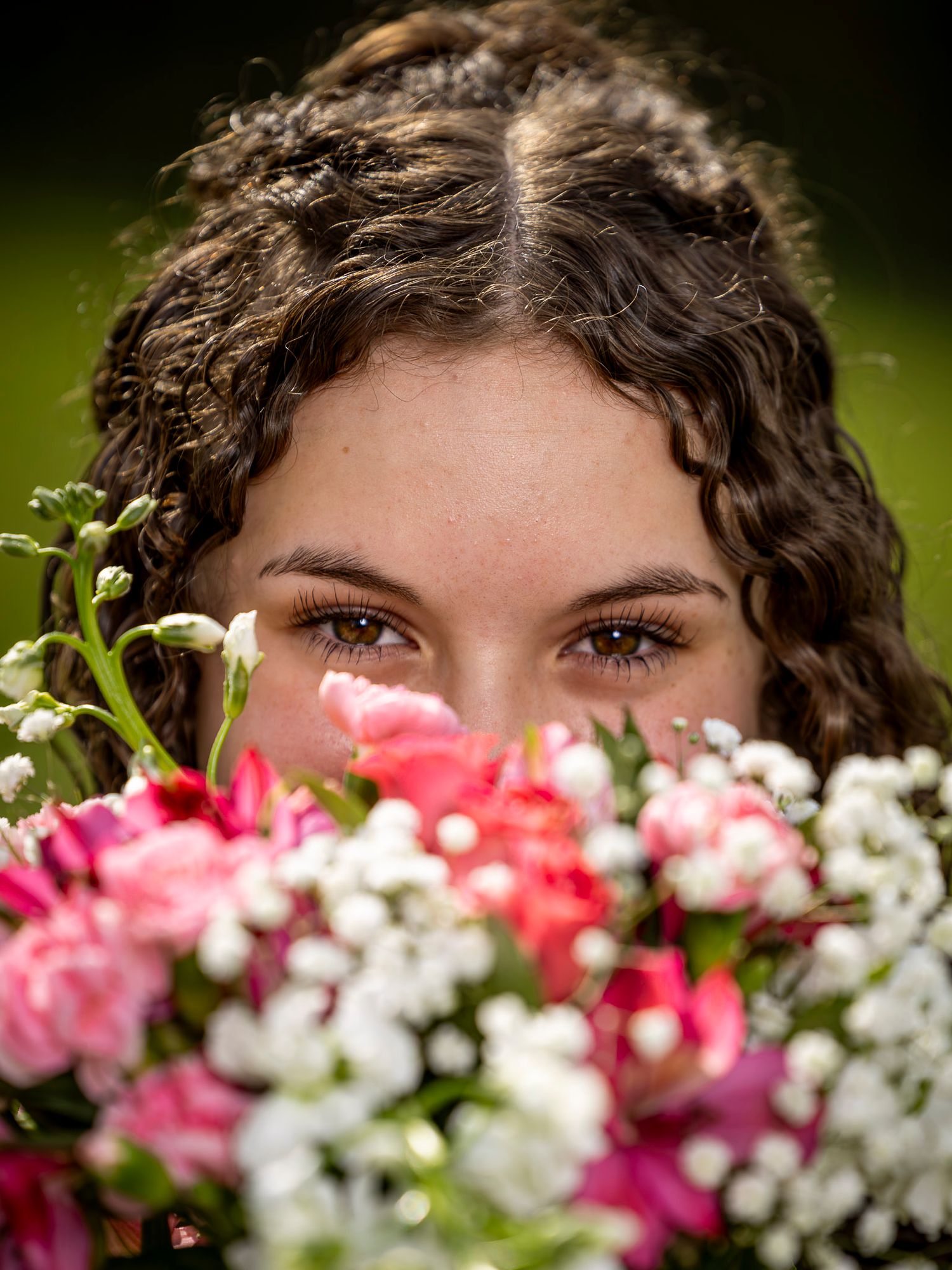 Young woman with curly brown hair peeks over a bouquet of pink and white flowers, showcasing her brown eyes.