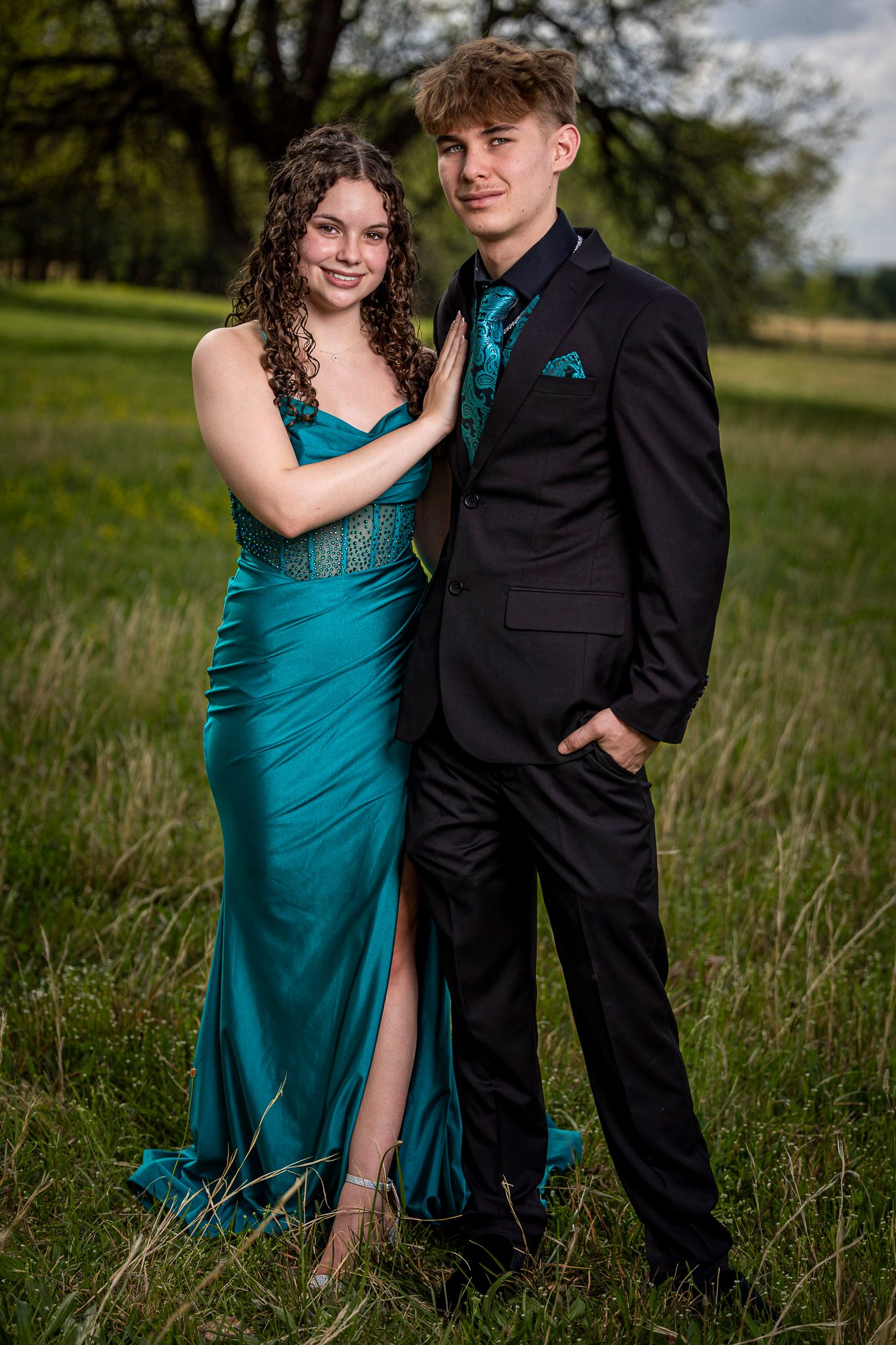 A couple poses in a green field wearing coordinating teal prom attire, she in a satin gown, he in a black suit.