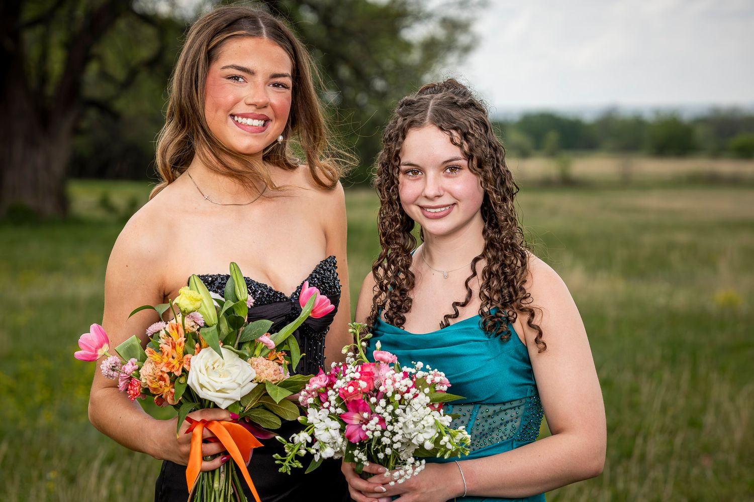 Two young women in formal dresses holding colorful bouquets outdoors in Prairie Grove Battlefield.