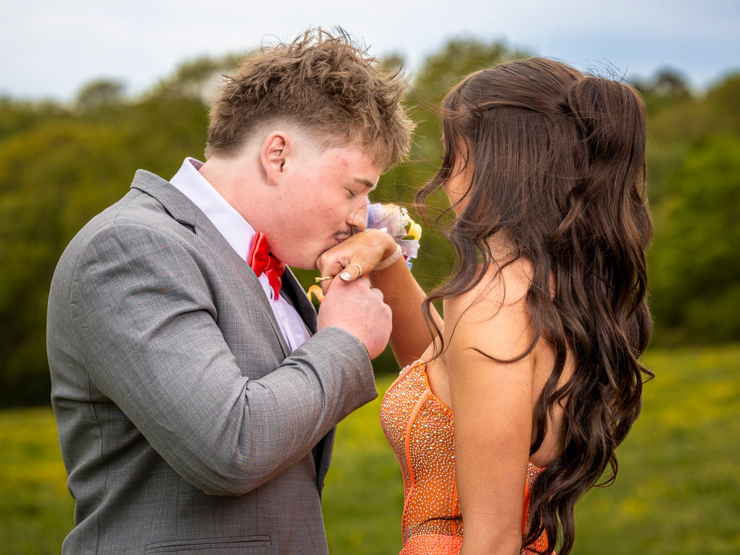 Young couple in formal wear, boy in gray suit kissing girl's hand in a green field at prom.