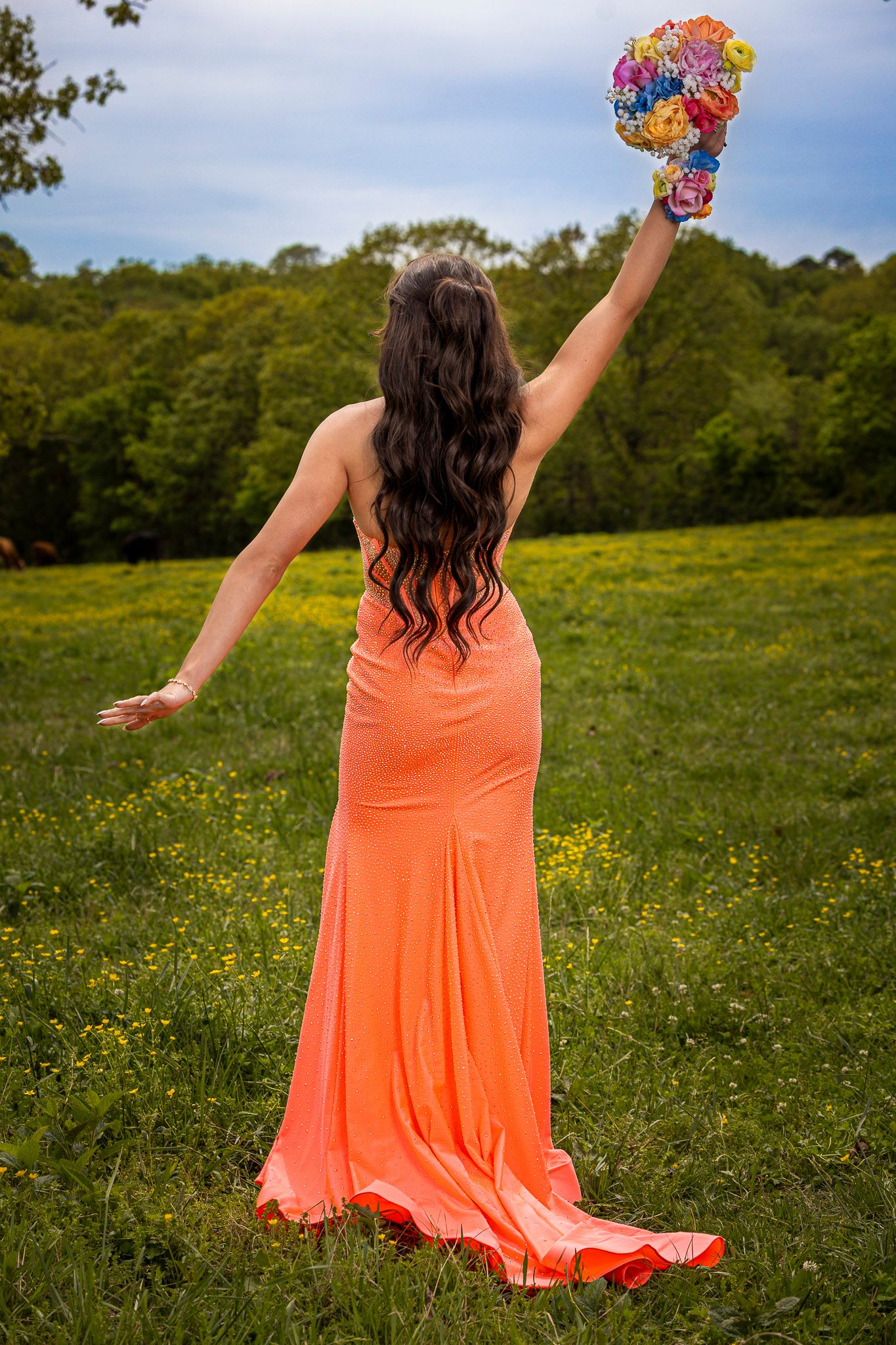 Woman in orange gown raising colorful bouquet in green meadow with wildflowers and trees in background.