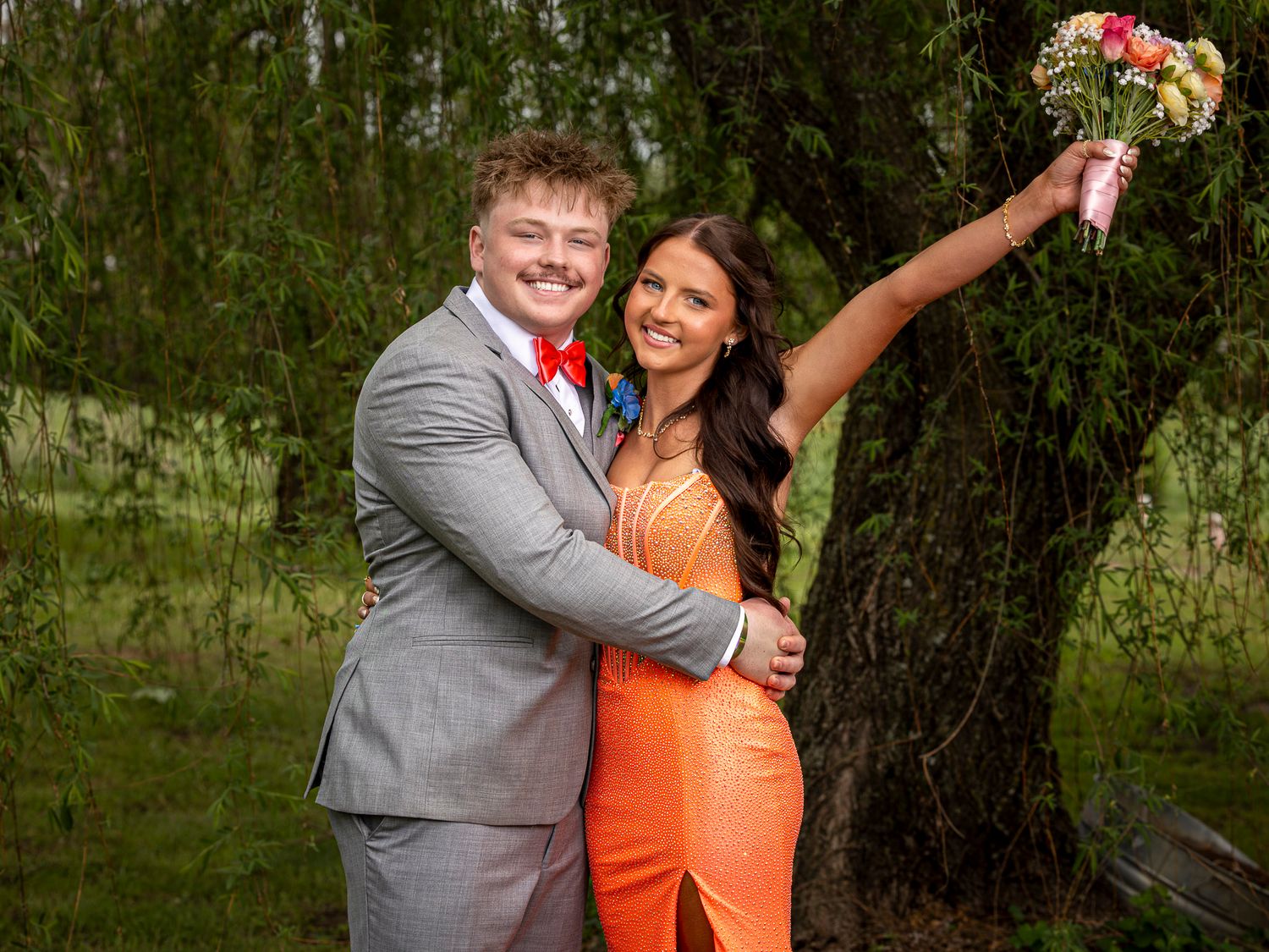 A smiling couple in prom attire, man in gray suit with red bow tie, woman in orange gown holding a floral bouquet.