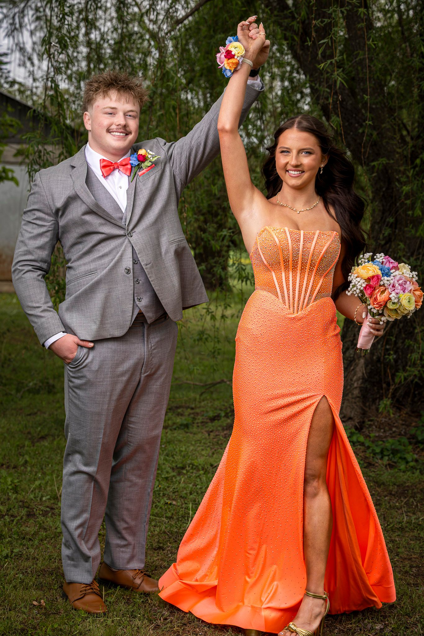 A couple in prom attire, woman in orange gown with bouquet, man in gray suit with bow tie, posing outdoors.